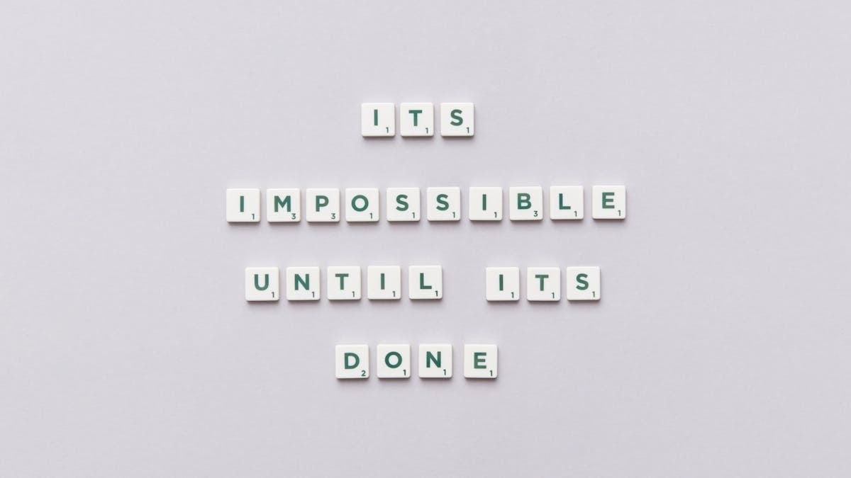 Motivational phrase 'It's impossible until it's done' arranged with Scrabble tiles on a gray background.
